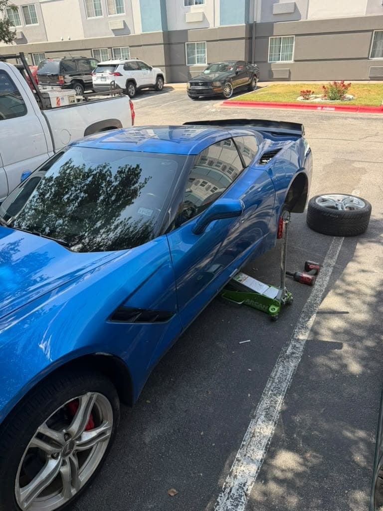 Chevrolet Corvette azul sobre un gato hidráulico en el estacionamiento de un hotel, con el rin trasero quitado y parado junto al carro.