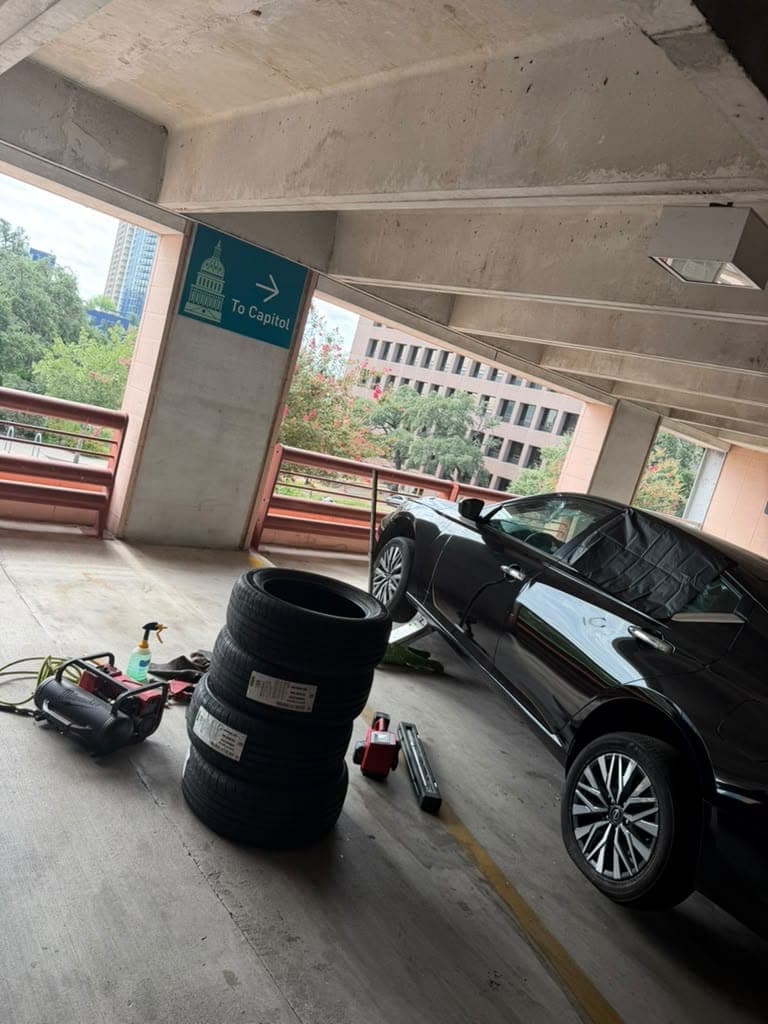 Four new tires stacked beside a black sedan in a downtown Austin parking garage, a sign on the column pointing to the Capitol.