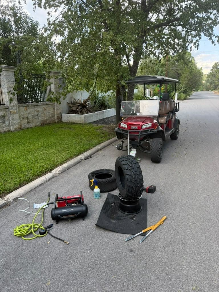 Carrito de golf Club Car rojo estacionado en una calle residencial, con una llanta de tacos parada sobre su rin, compresor y herramientas en el pavimento.