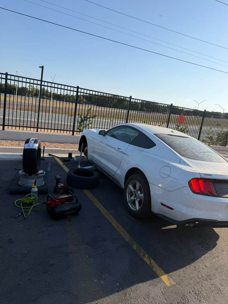 White Ford Mustang on a jack beside a highway fence, a new tire being fitted to its wheel on the ground, compressor and impact wrench alongside.