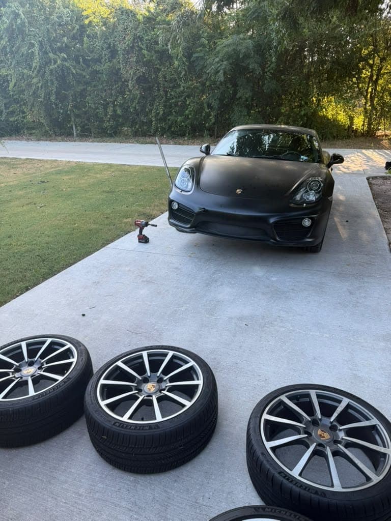 Matte black Porsche Cayman parked on a driveway with three new Michelin tires on their wheels lined up in front of it.