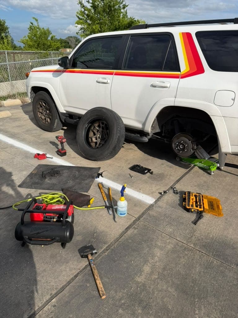 White Toyota 4Runner with retro stripes on a jack in a parking lot, its rear wheel and tire lying beside it and tools on the ground.