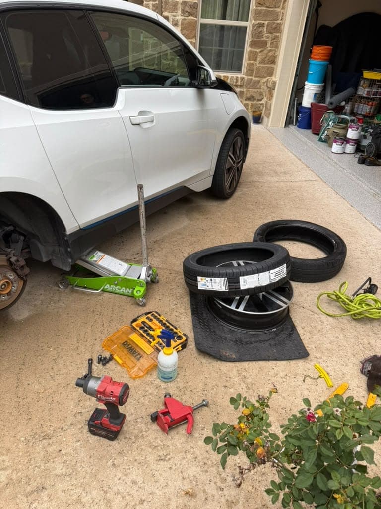White BMW i3 on a jack in a driveway, a new tire still wearing its label mounted on its wheel, the worn old tire beside it.