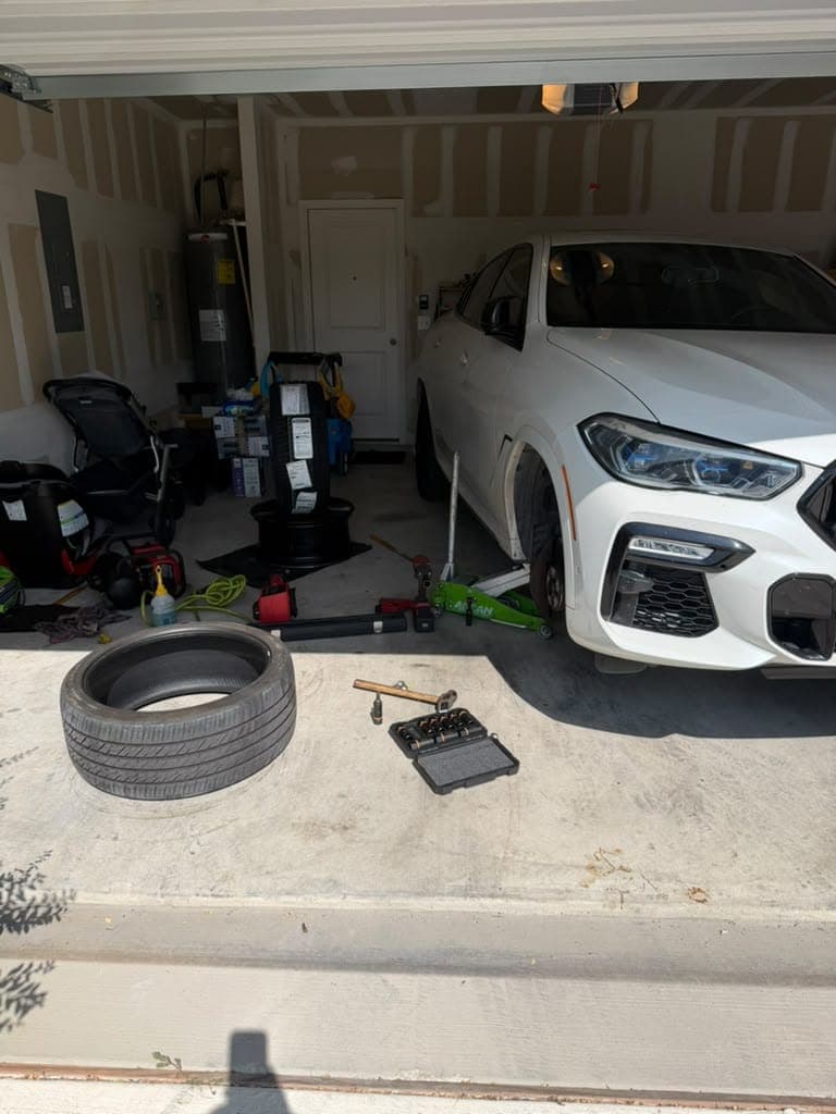 White BMW X6 on a floor jack inside a home garage, front wheel removed, a socket set and a tire on the floor.