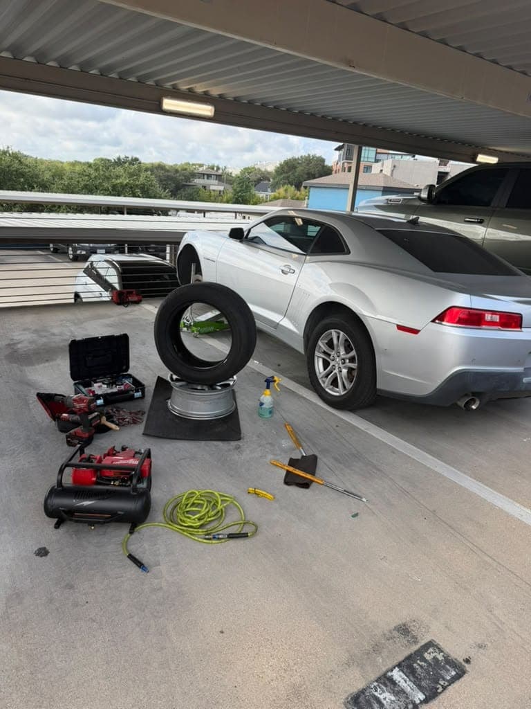 Silver Chevrolet Camaro in a covered parking deck with a tire dismounted from its wheel on a mat, tools and a compressor nearby.