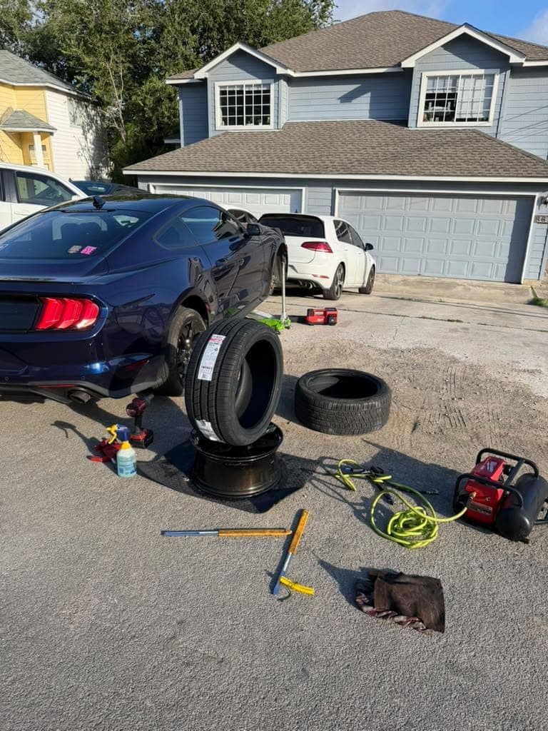 Dark blue Ford Mustang in a driveway with a new tire mounted on a black wheel standing beside the car and the old tire behind it.